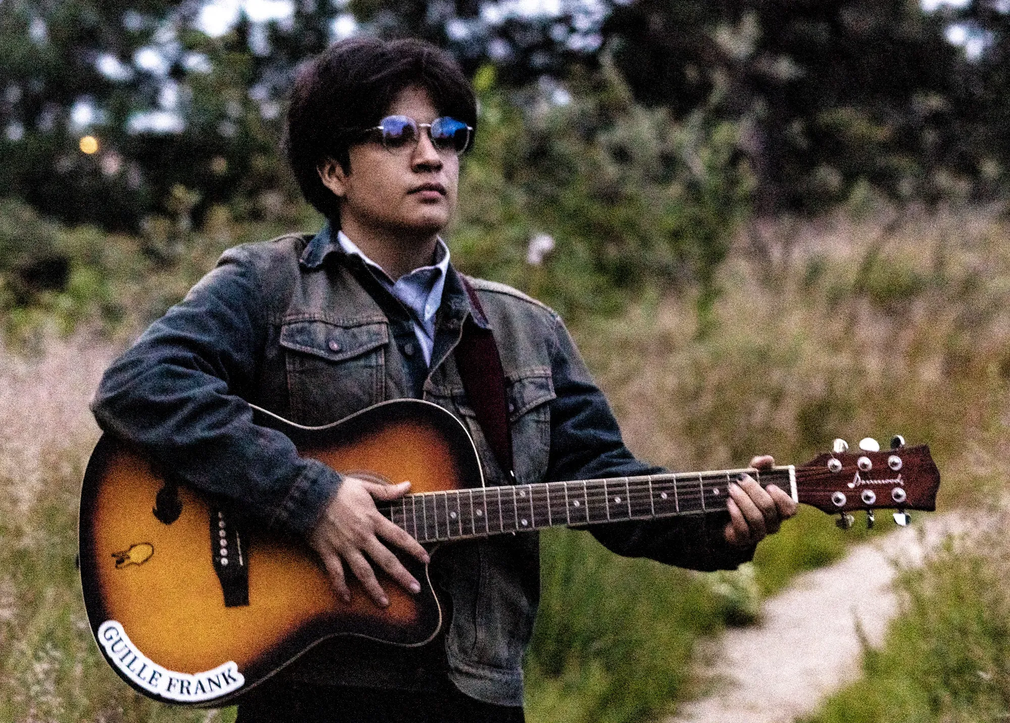 Guille Frank con lentes azules y su guitarra acústica en un sendero